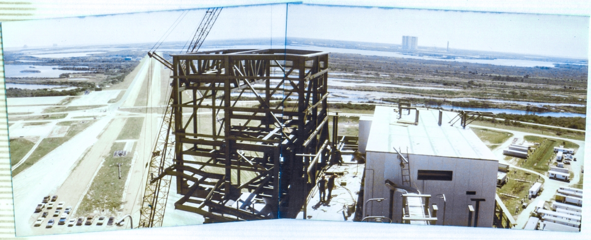 Viewed from a vantage point on the Fixed Service Structure at Space Shuttle Launch Complex 39-B, Kennedy Space Center, Florida, 240’ above the wilderness around it, Union Ironworkers from Local 808, working for Wilhoit Steel Erectors assemble structural framing which will become the Reaction Control System Room, cantilevered forward, above the main body of the Rotating Service Structure, which is out of frame in this image, beneath the RCS Room. To the right of the RCS Room, the Hoist Equipment Room, which houses the 90-ton Payload Canister Hoist, gleams white with its skin of newly-installed insulated-metal paneling. In the far distance to the right, the Vertical Assembly Building and the last remaining Launch Umbilical Tower from the Apollo Program stand above the horizon.